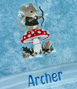 Close-up of a light blue towel featuring an embroidered mouse archer on a red-and-white mushroom, with the name 'Archer' embroidered in blue at the bottom.